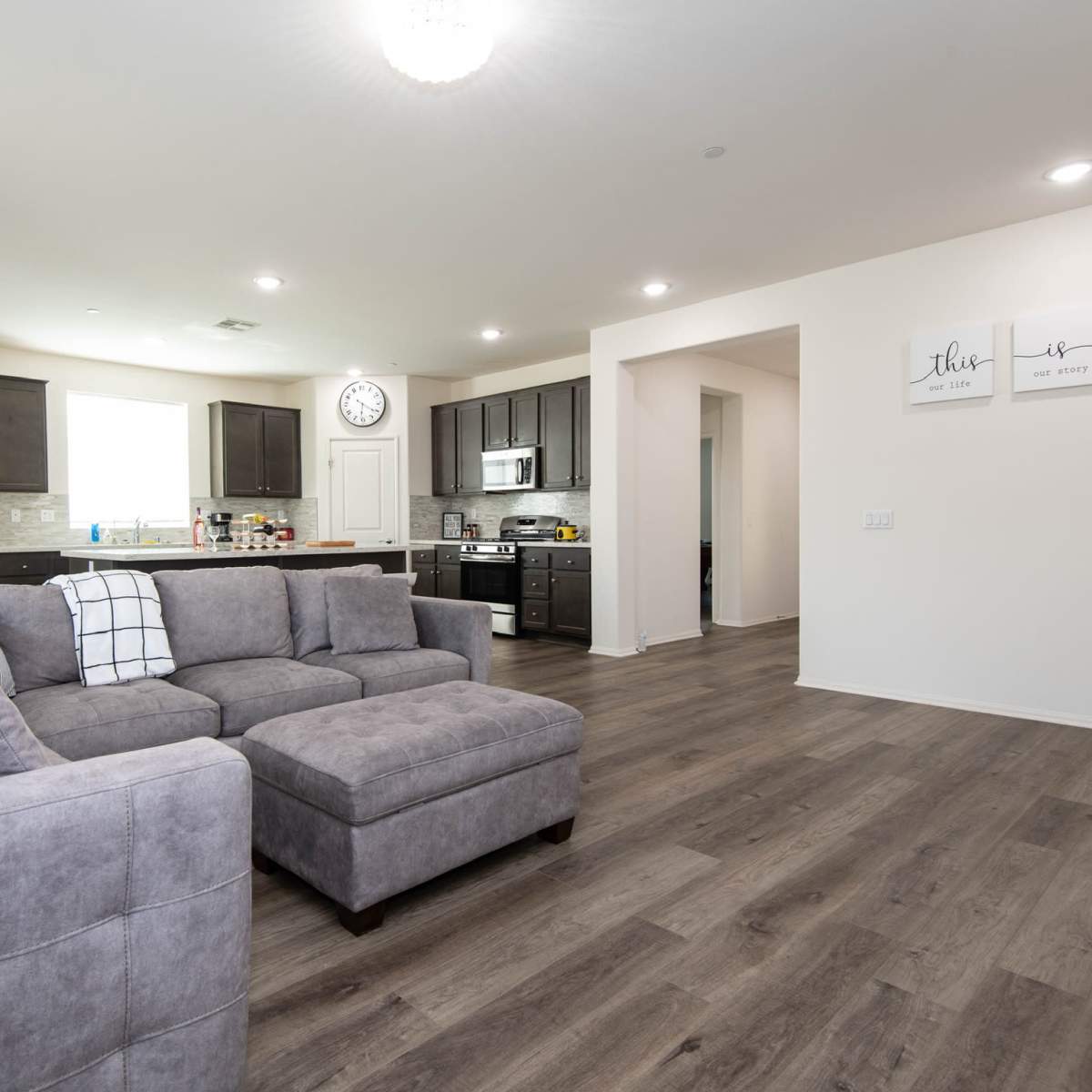 A modern living room features a gray sectional sofa, hardwood floors, and an open kitchen with dark cabinets and stainless steel appliances.