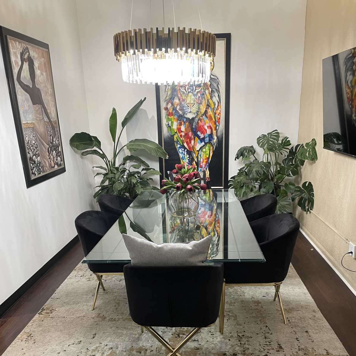 A modern dining room with a glass table surrounded by black velvet chairs, a large colorful lion painting on the wall, and indoor plants on either side.
