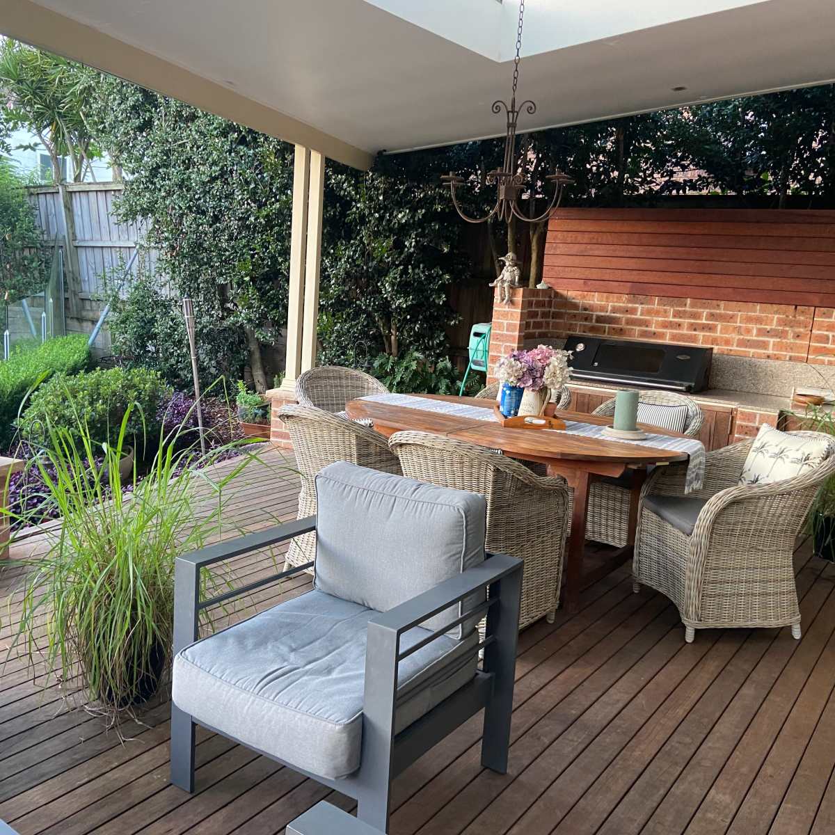 A spacious outdoor patio features a wooden dining table surrounded by wicker chairs, alongside potted plants and a barbecue grill under a covered roof.