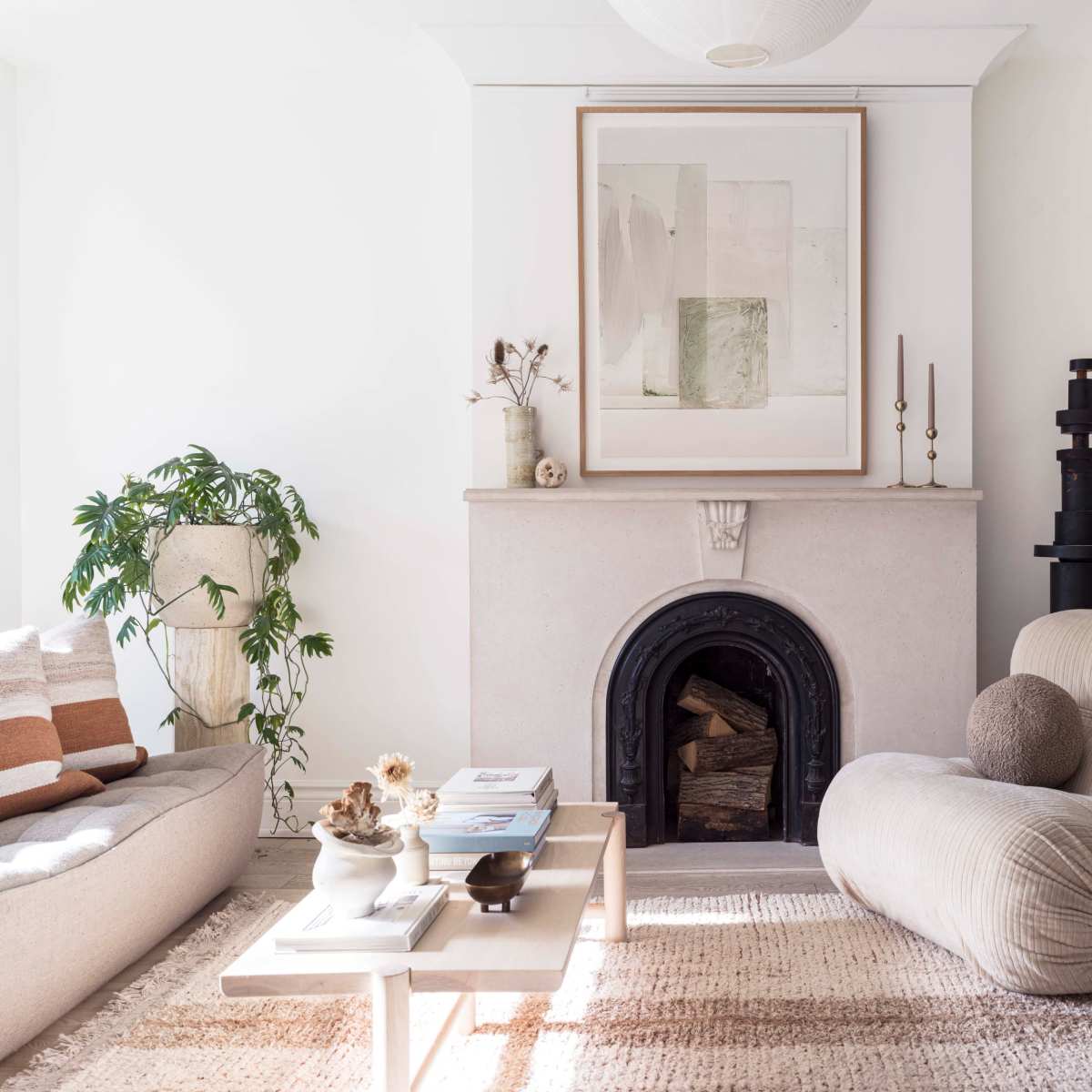 A bright living room features a fireplace with a framed artwork above it, a sofa with decorative pillows, a coffee table with books, and a wooden stool beside a soft, sculptural chair.