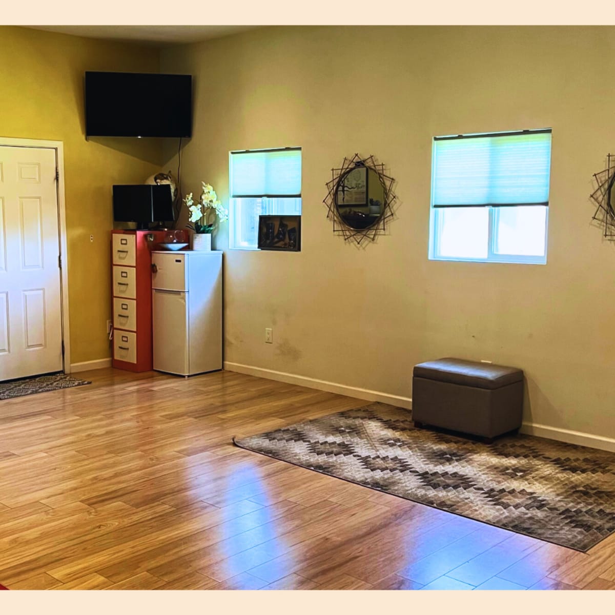 A spacious interior room with wooden flooring, featuring a door, two windows, a television mounted on the wall, and various furnishings including a bench and storage units.