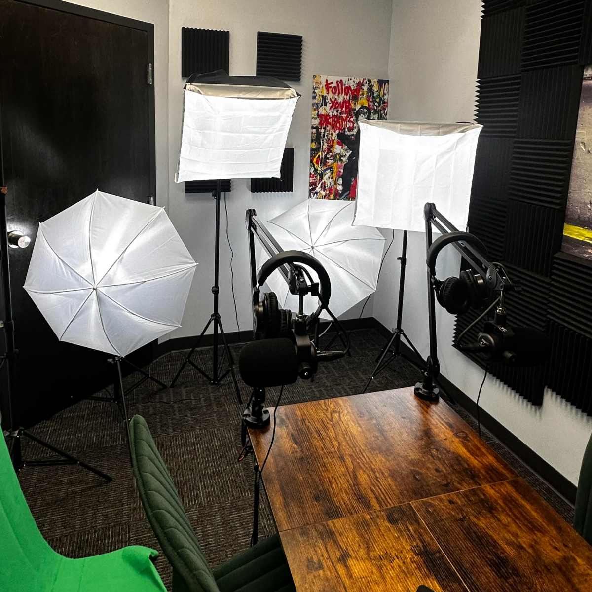 A small recording studio with two tripods holding softbox lights, two microphones on stands, and a table surrounded by various soundproofing panels on the walls.