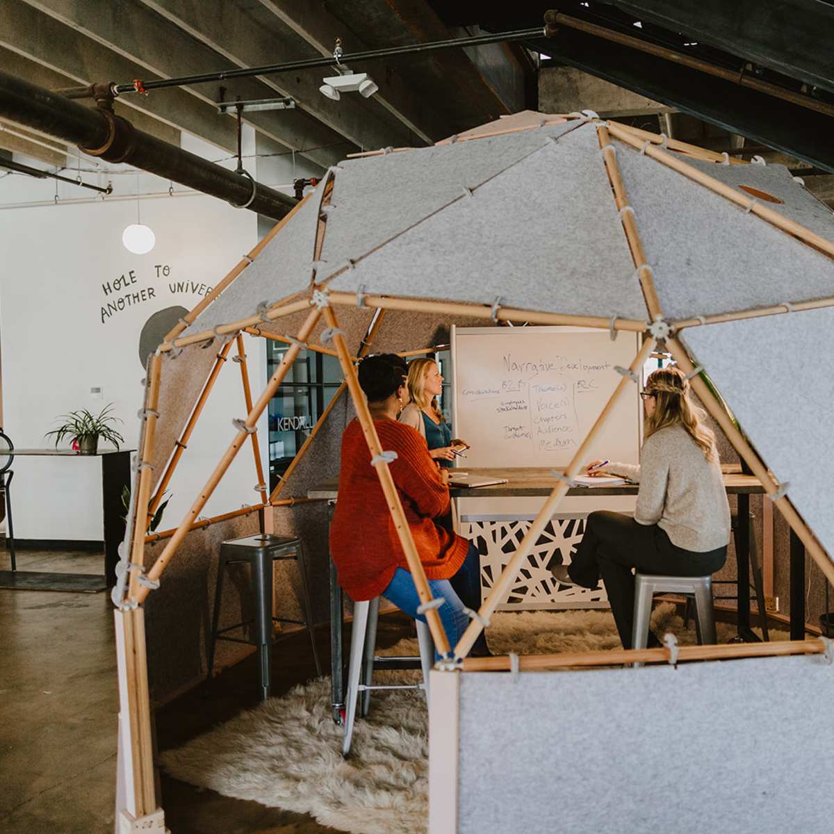 Two people are sitting at a round table inside a geometric, fabric-covered structure in a modern office space, engaged in a discussion with notes on a whiteboard nearby.
