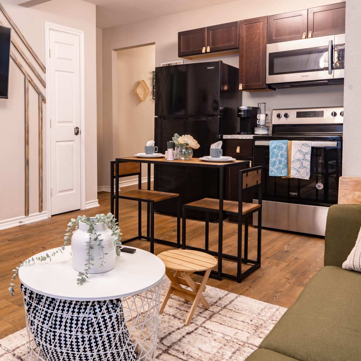 A modern living space featuring a green sofa, a round, woven coffee table, a small dining area with two stools, and a kitchen with dark cabinets and stainless-steel appliances.