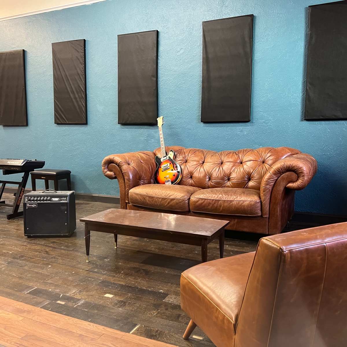 A music practice room with a brown leather sofa, a guitar resting on it, an amplifier, a small table, and a keyboard set against a blue wall with soundproofing panels.