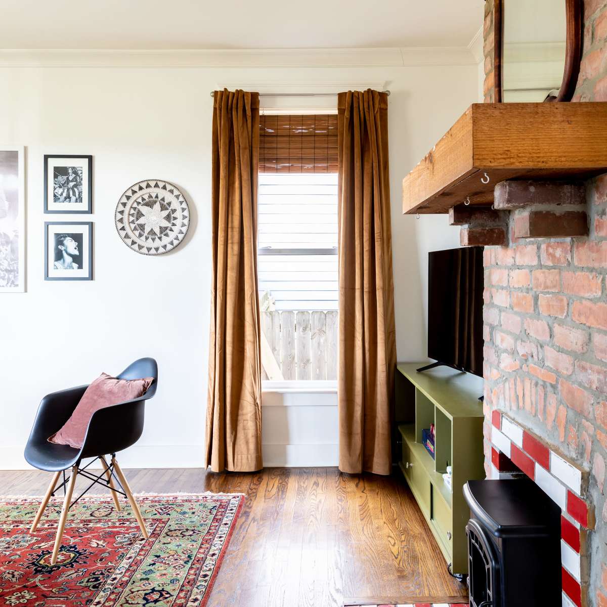 A living room features a brick fireplace, a television on an entertainment unit, framed artwork on the walls, and a window with brown curtains.