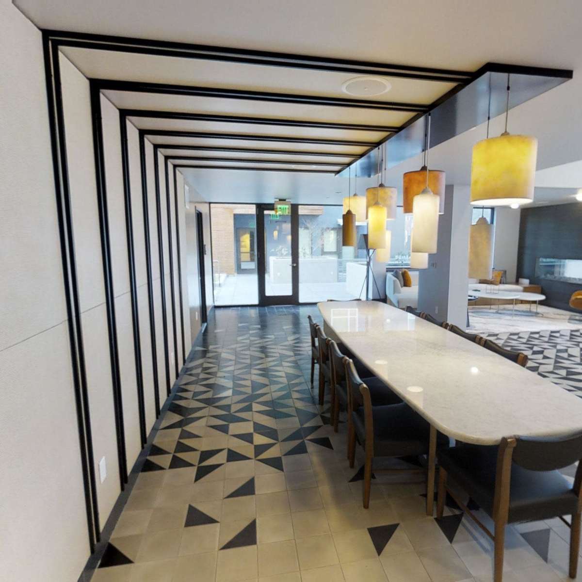 A modern interior space featuring a long table surrounded by chairs, decorative pendant lights, and a patterned floor design.