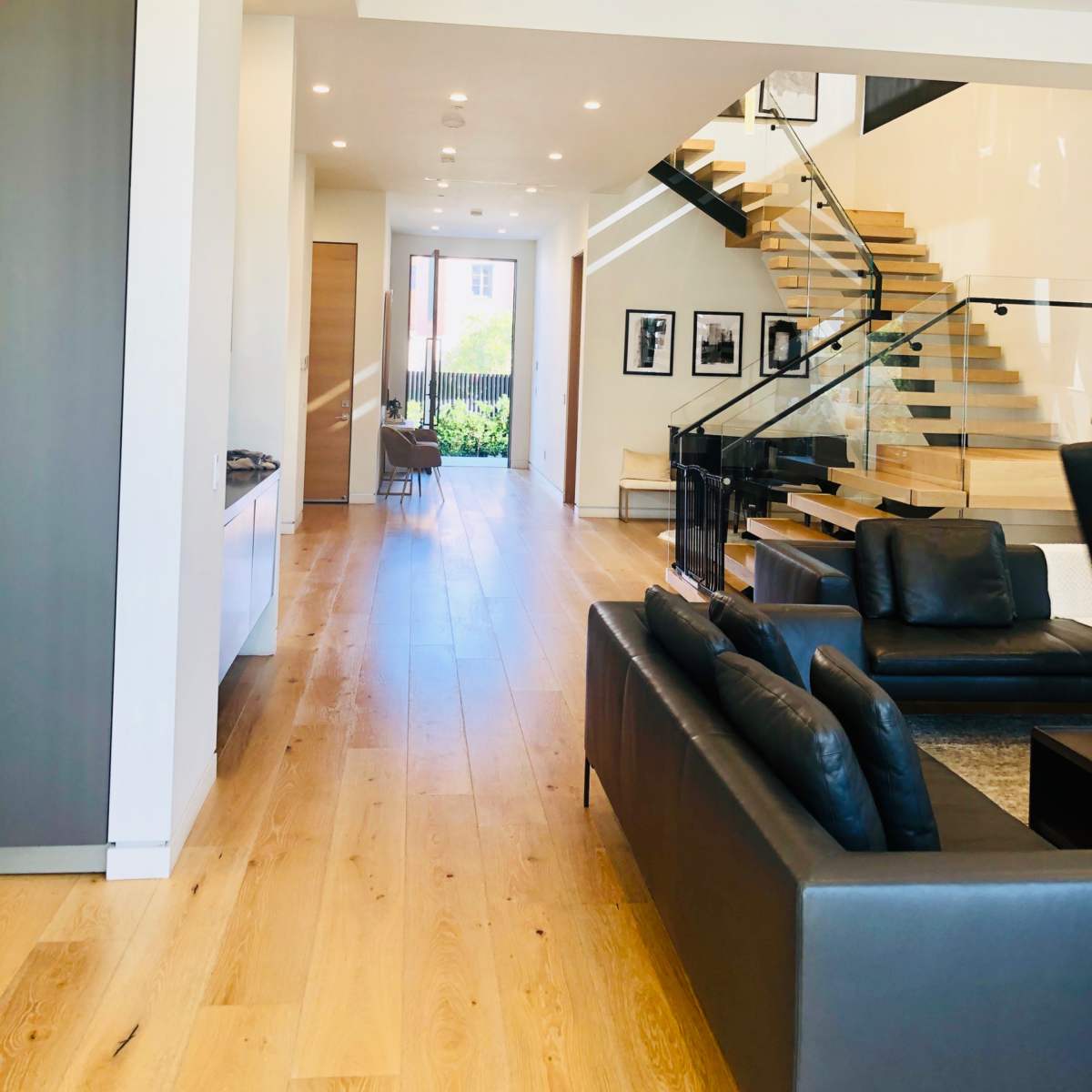 A modern interior of a house featuring a long hallway, a staircase, and a living area with a black couch.