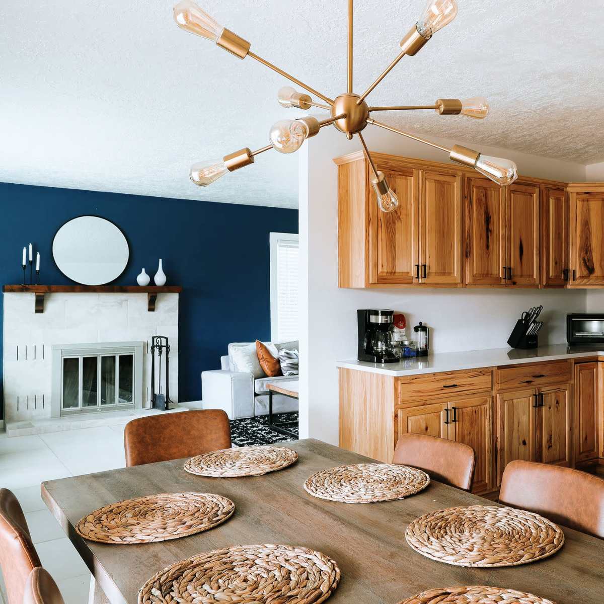 A modern kitchen features wood cabinetry, stainless steel appliances, a dining table set with woven placemats, and a blue accent wall.