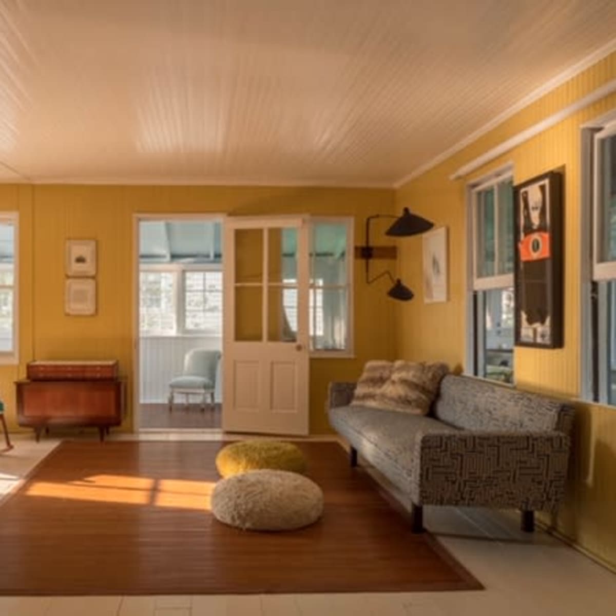 A bright, yellow-walled room featuring a sofa, armchair, a wooden side table, and large windows letting in natural light.