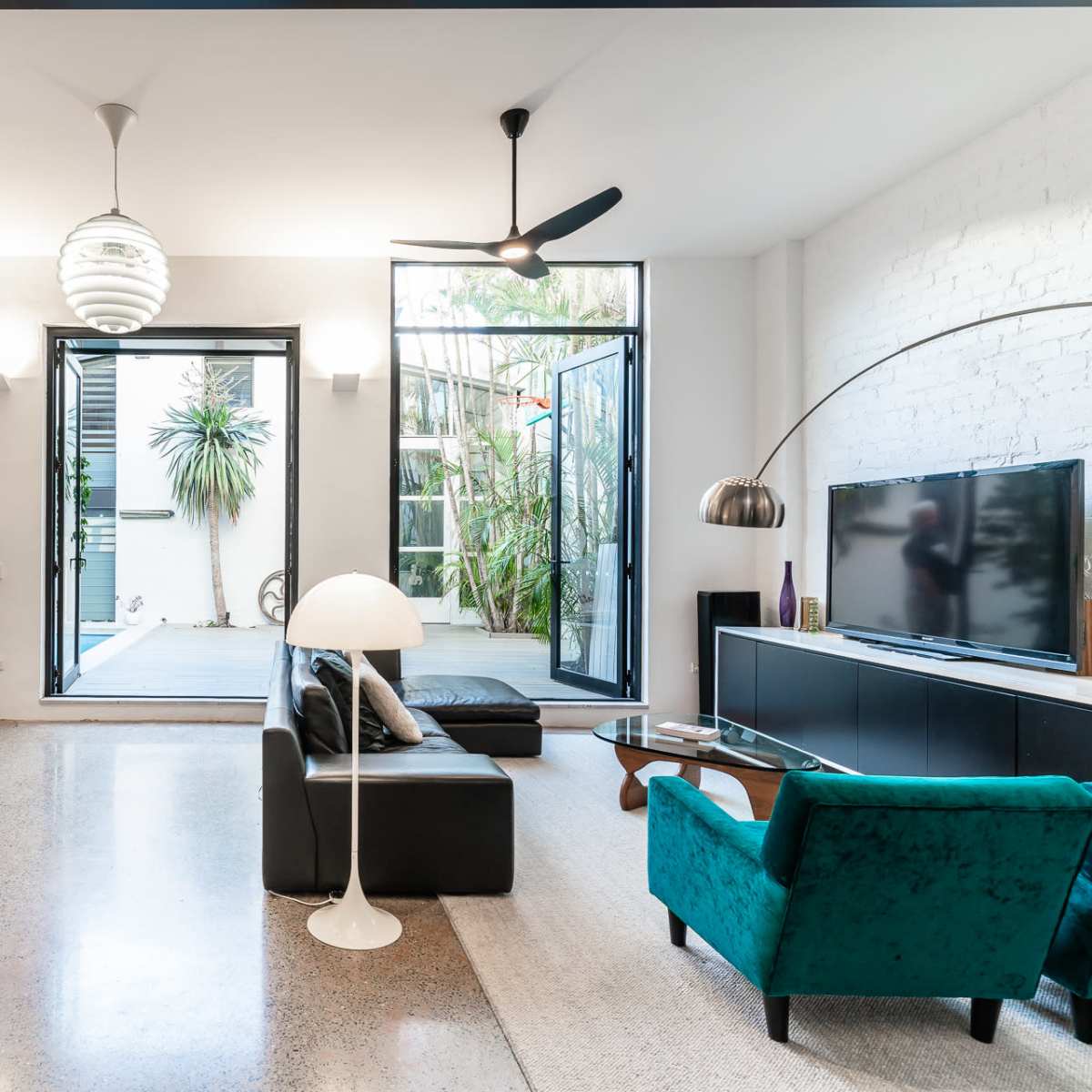 A modern living room featuring a black leather sofa, a green armchair, a large television, and glass doors opening to a patio with plants.