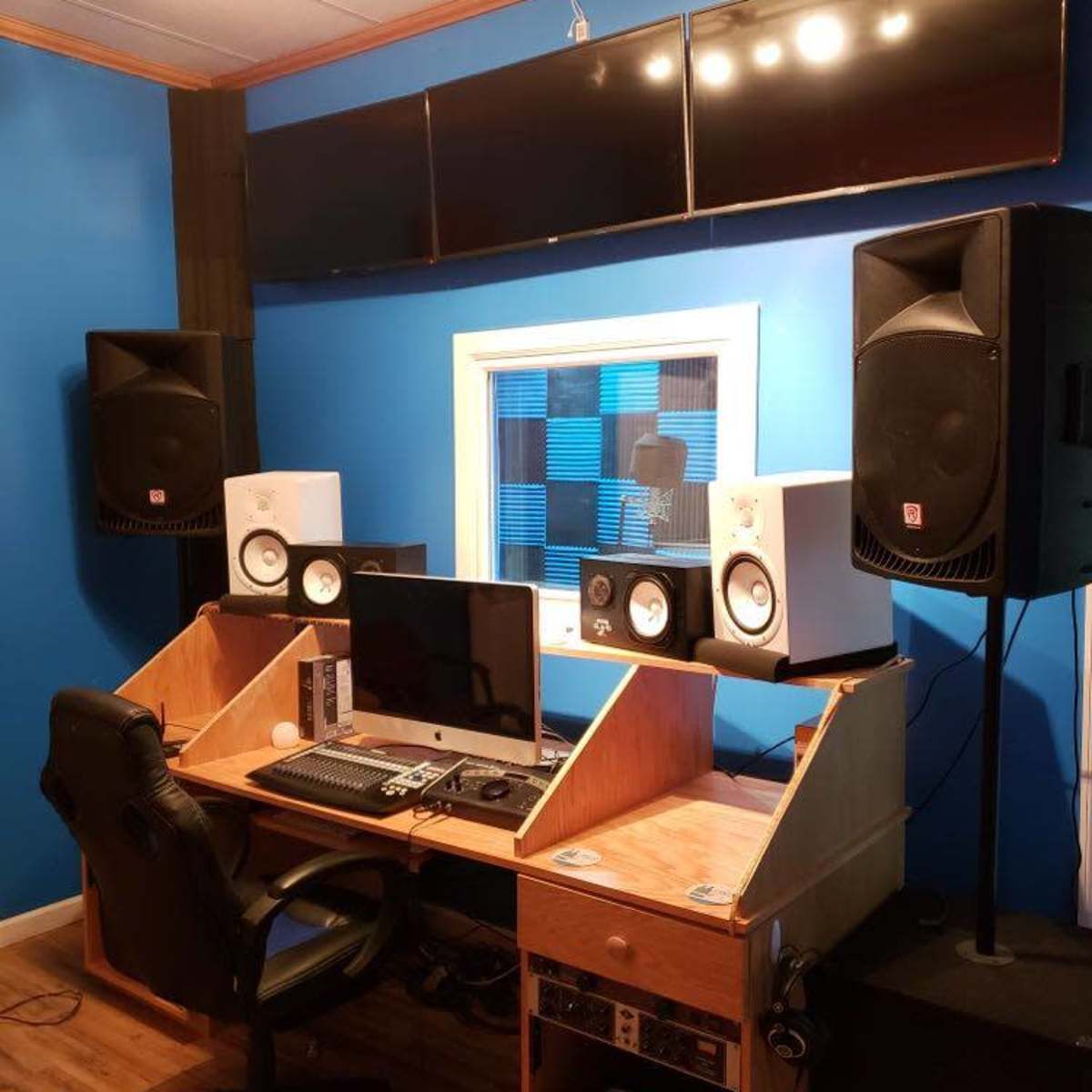 A music studio with a computer desk, audio monitors, and multiple screens mounted on a blue wall.