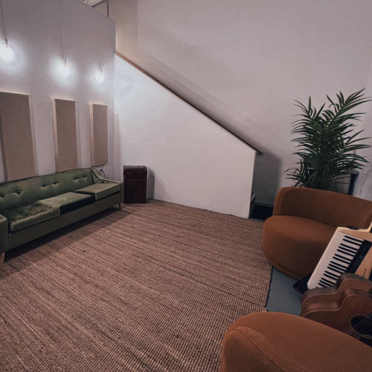 A cozy music studio featuring a green velvet sofa, an orange chair, a staircase, and soundproof panels on the walls.