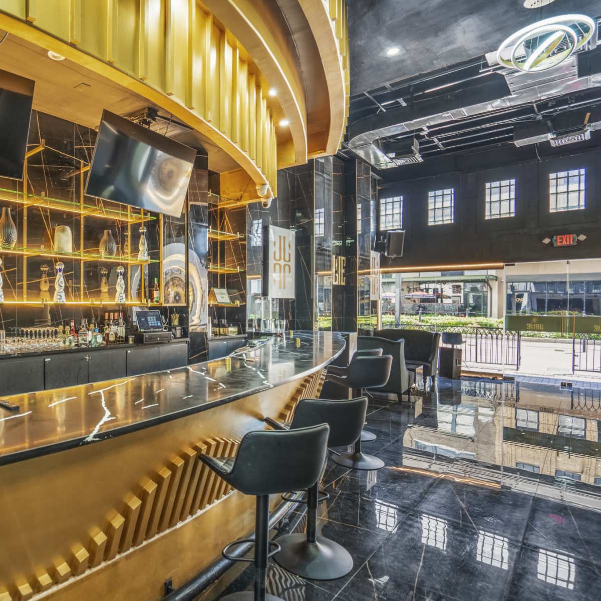 A modern bar area with a sleek curved counter, illuminated shelves featuring a variety of liquor bottles, and large wall-mounted screens.