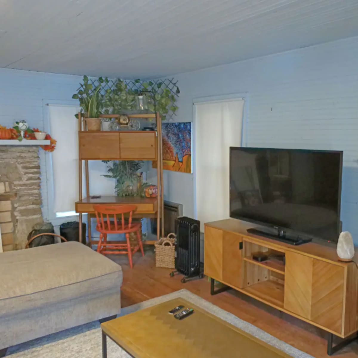 A living room features a sectional sofa, a wooden TV stand with a large television, a desk with a red chair, and decorative plants on shelves against a light blue wall.