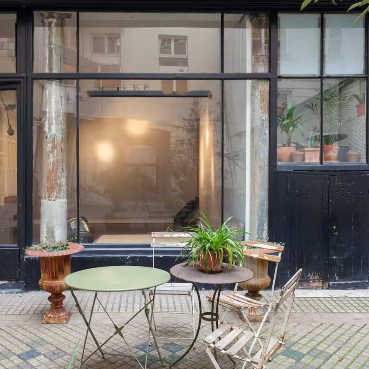 A small outdoor seating area with a green table and two metal chairs is set in front of a large glass storefront featuring plants and minimal interior decor.