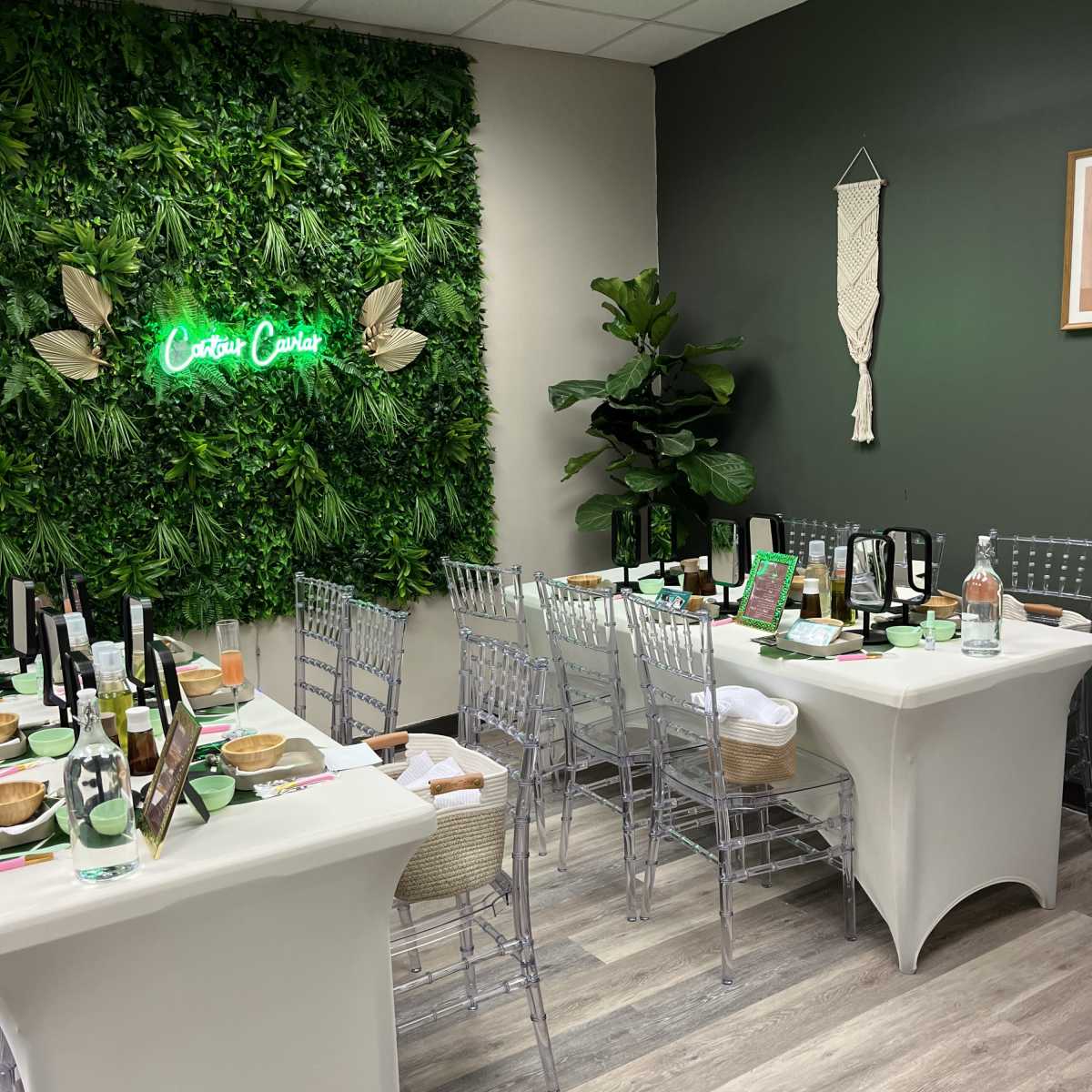 A dining setup with clear chairs, a green foliage wall, and a neon sign, arranged for a gathering.