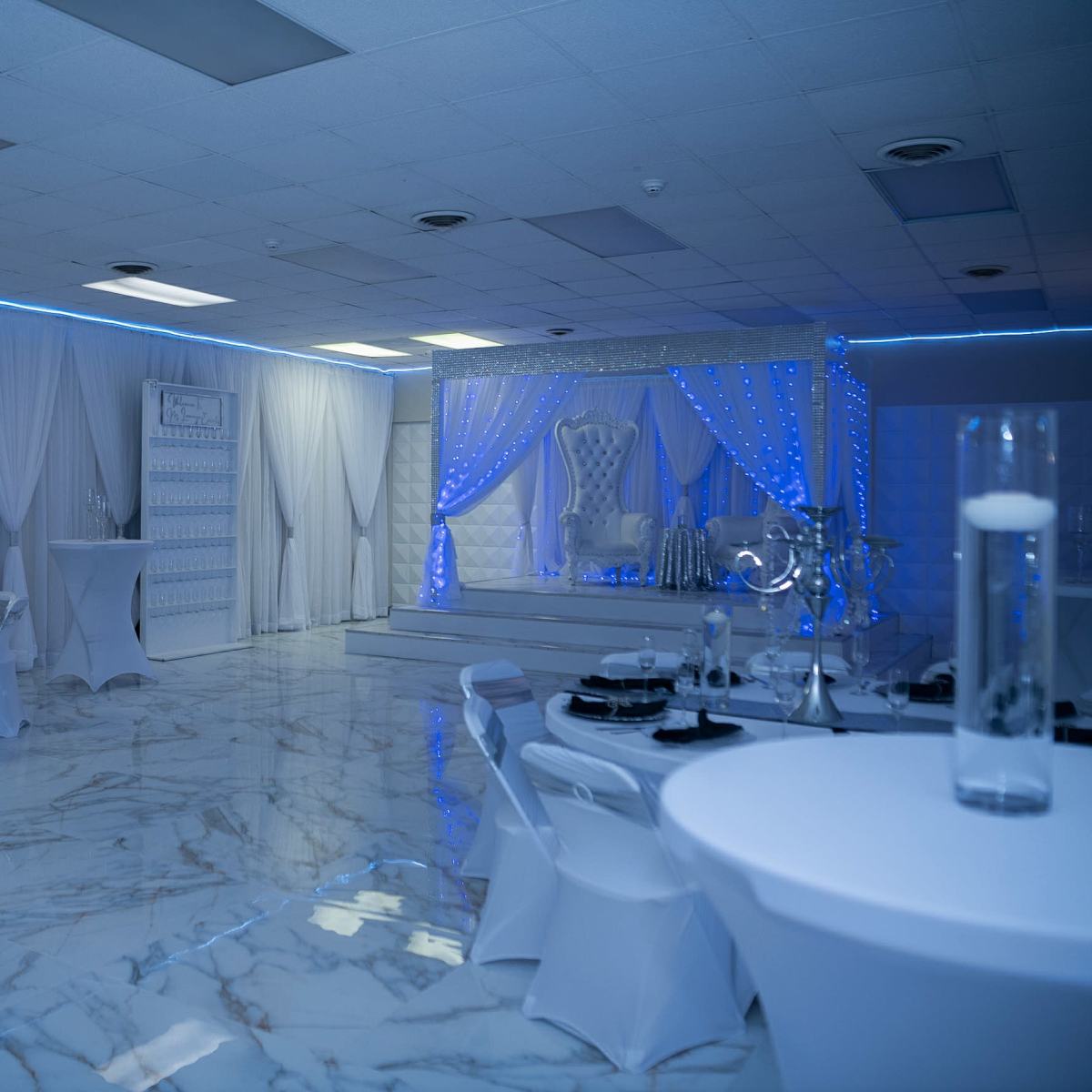 A spacious event hall features white table settings, elegant decor, and blue lighting, with a stage at the front adorned with drapes and a decorative throne.