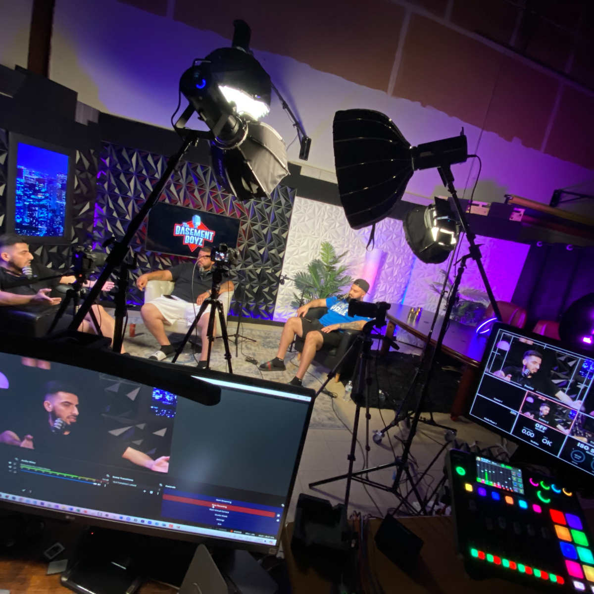 A recording studio setup with three individuals sitting on chairs, surrounded by cameras, lights, and a production console displaying live feeds.