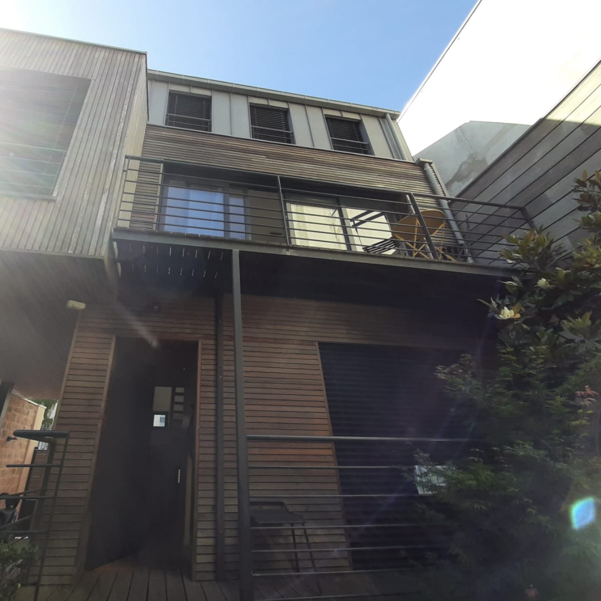 A two-story wooden structure with a balcony and a door at the ground level, surrounded by greenery under a clear blue sky.