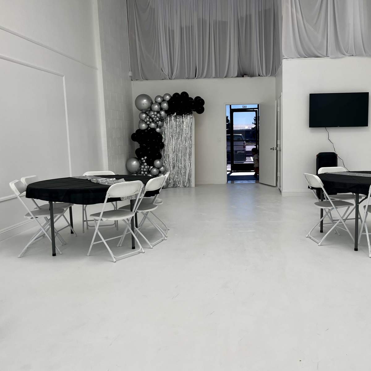 A spacious, white-walled room with black and white tables set for an event, featuring a decoration of black and silver balloons in one corner and a television mounted on the wall.