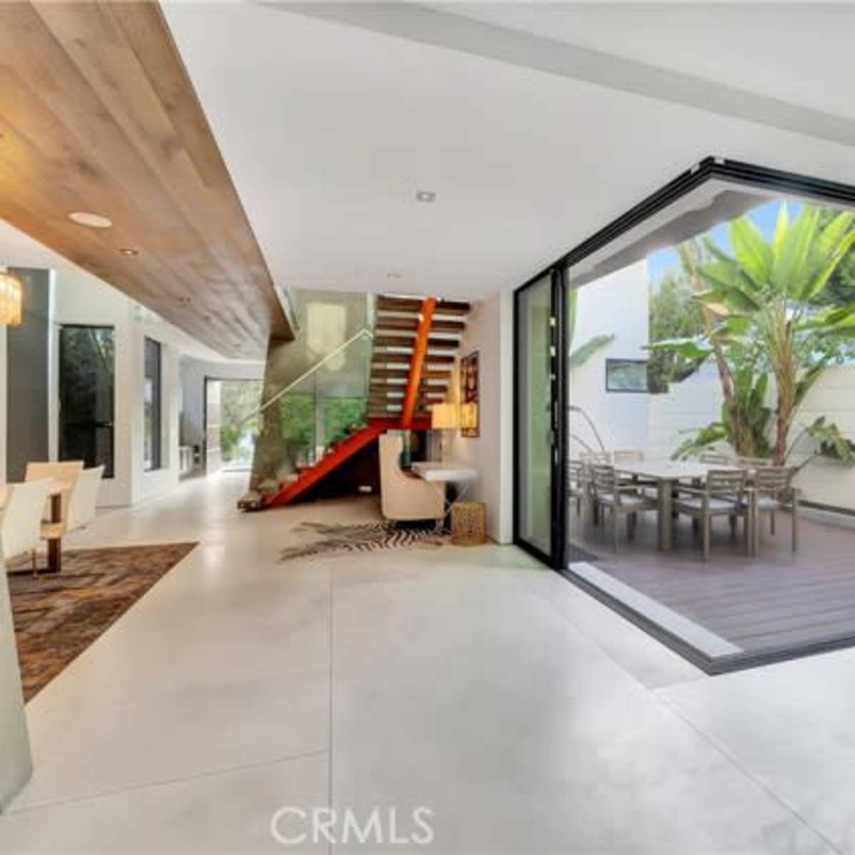 A modern interior space featuring a wooden staircase, large glass doors opening to an outdoor area, and a dining table surrounded by greenery.