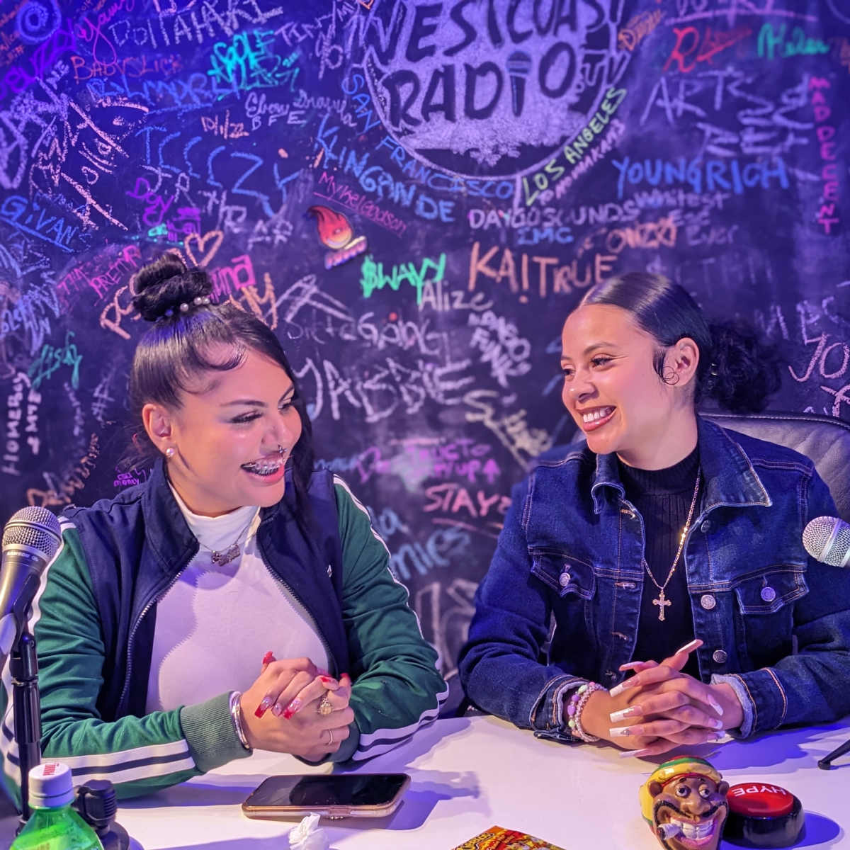 Two women sit at a table, smiling and conversing in front of a colorful graffiti-covered wall.