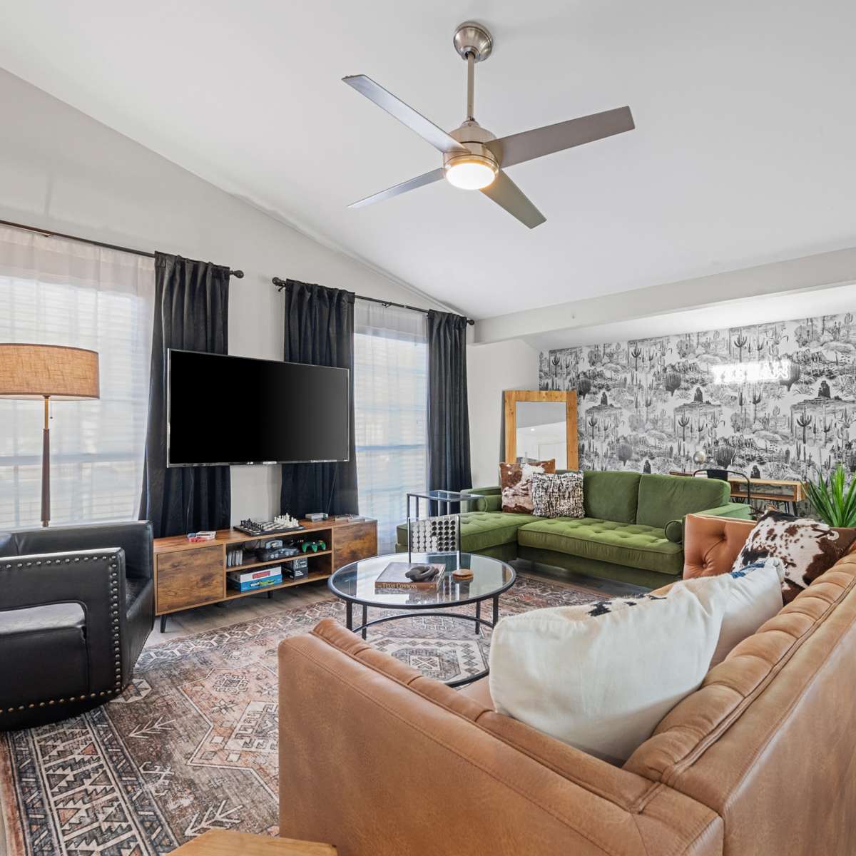 A modern living room features a green sectional sofa, a brown leather couch, a coffee table, and a large wall-mounted television against a patterned wallpaper backdrop.