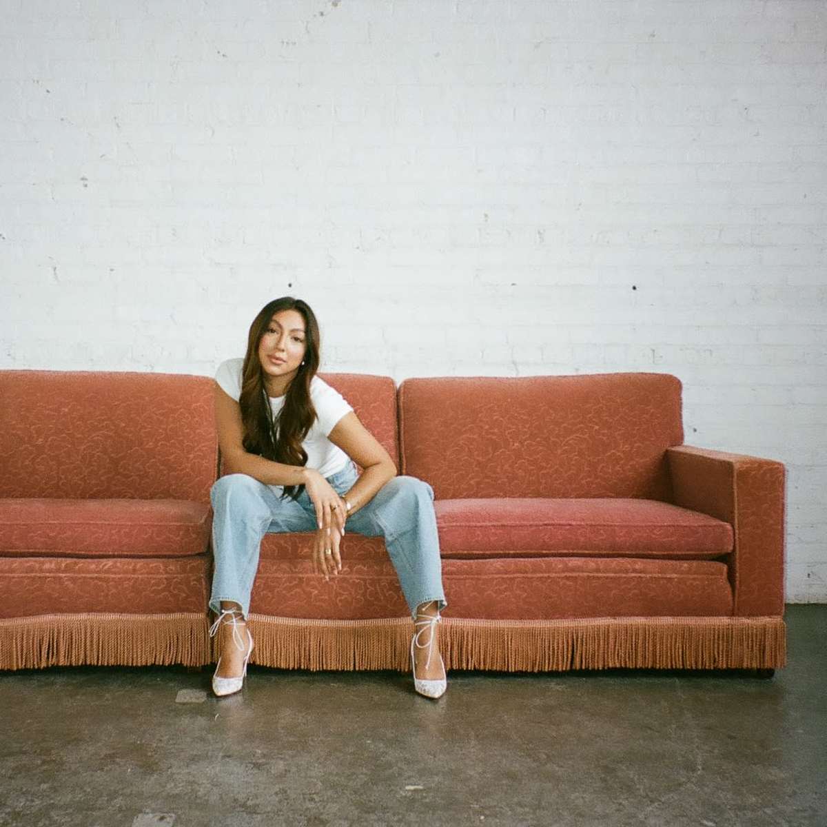 A woman sits on a vintage red couch against a white brick wall.