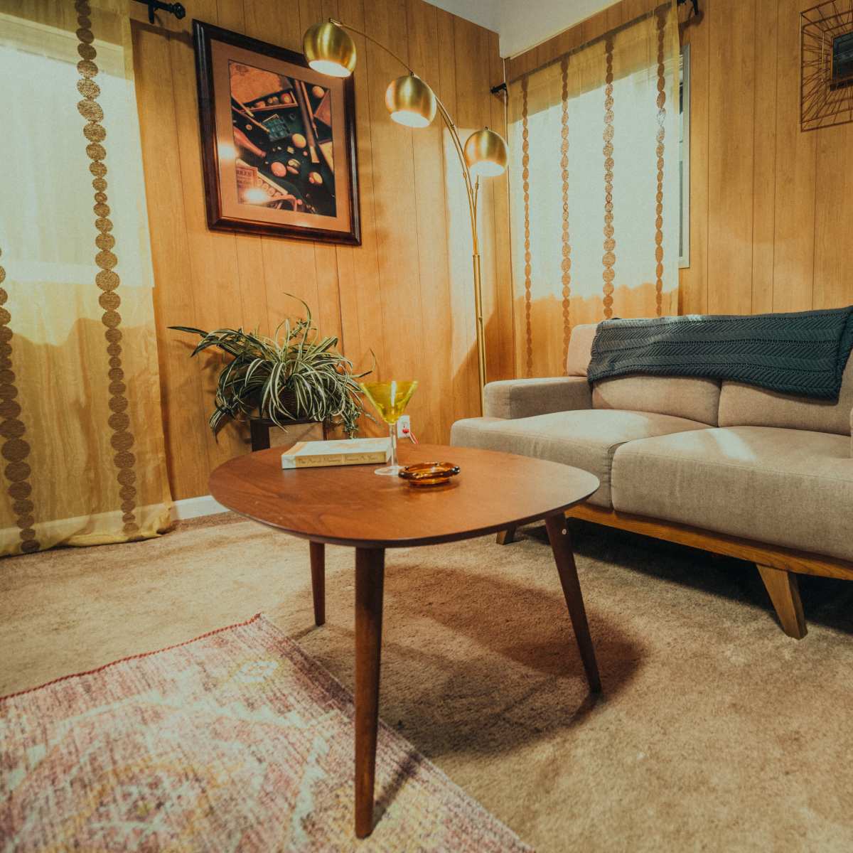 A cozy living room with a gray sofa, a wooden coffee table, a lamp, and decorative curtains against a paneled wall.