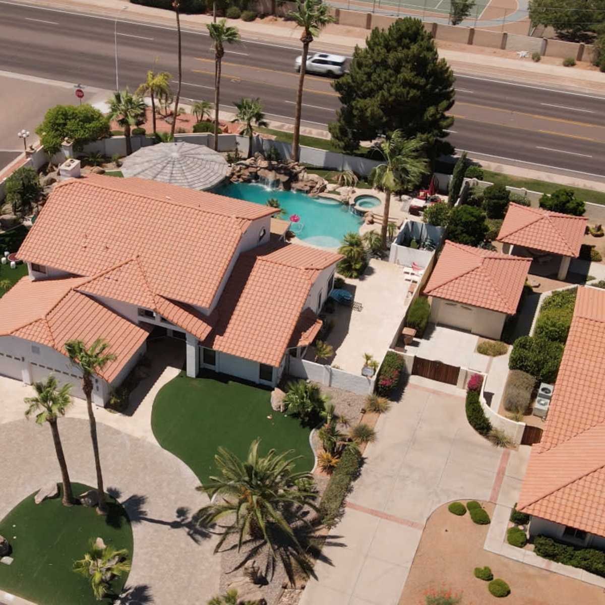 An aerial view of a suburban home surrounded by neatly landscaped lawns and palm trees, with a pool in the backyard and a road nearby.