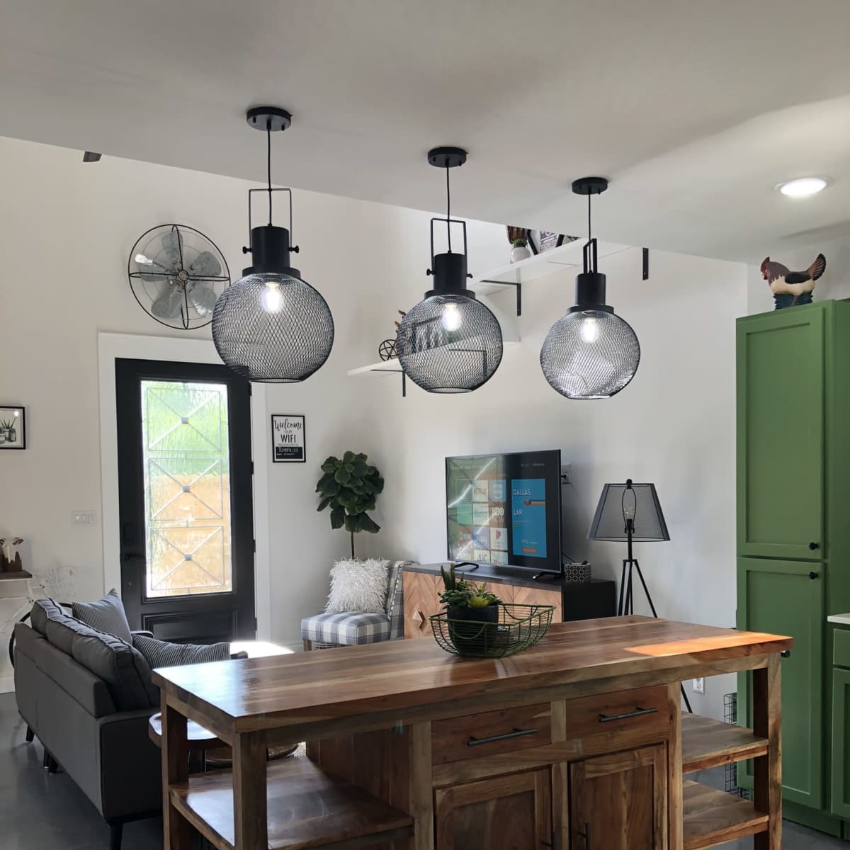A wooden kitchen island with shelves stands in a modern living space featuring a gray sofa, a television, and pendant lights above.