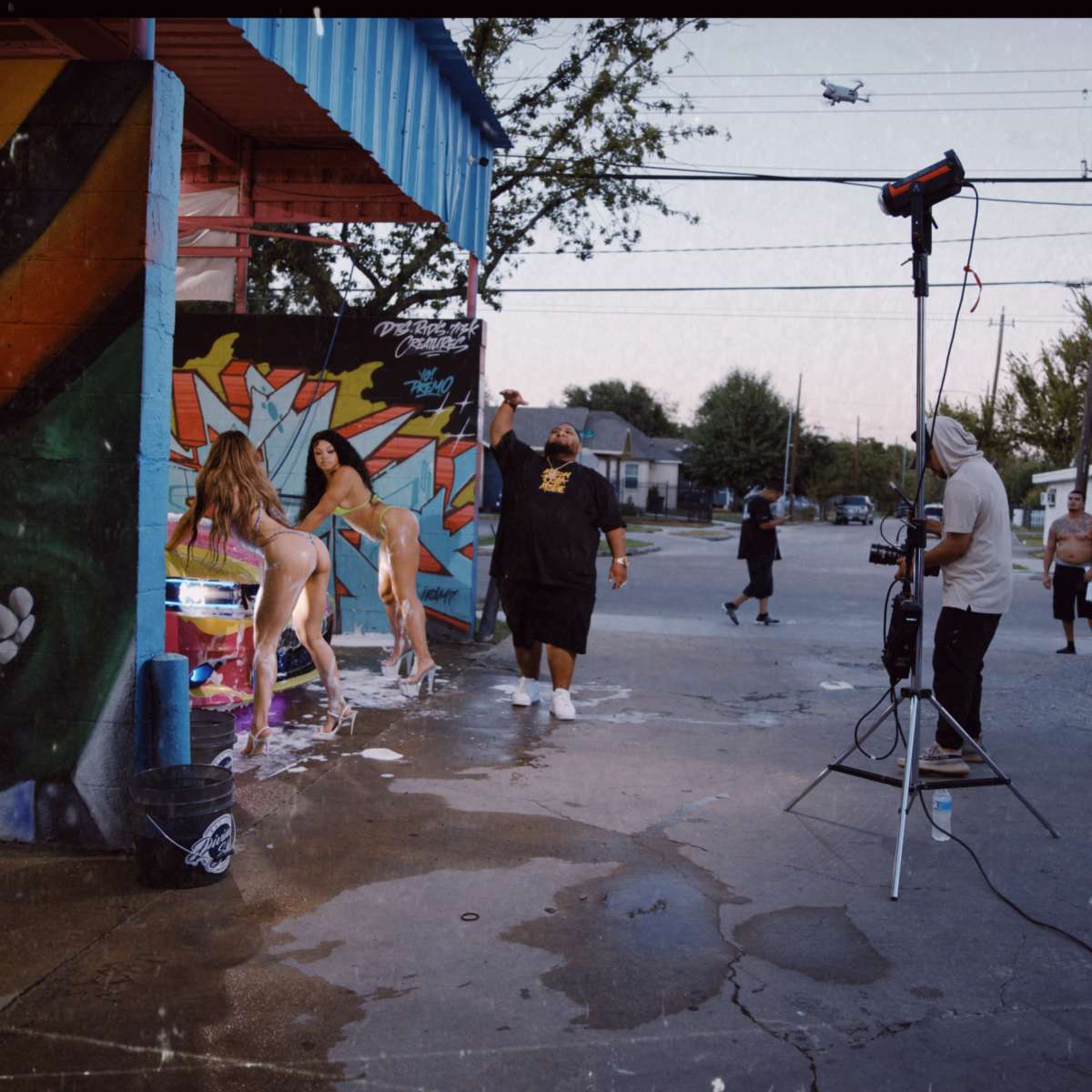 A group of people are engaged in a photoshoot outside a colorful mural, with one person posing in water while others prepare equipment and watch.