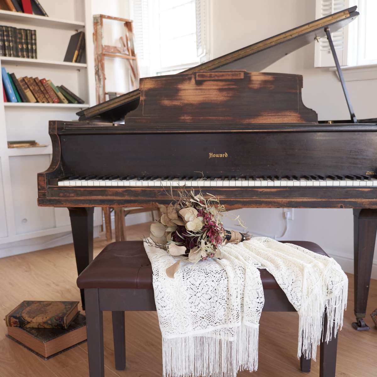 A vintage piano is placed next to a wooden bench, adorned with a lace cloth and a dried flower bouquet, in a room featuring bookshelves and natural light.