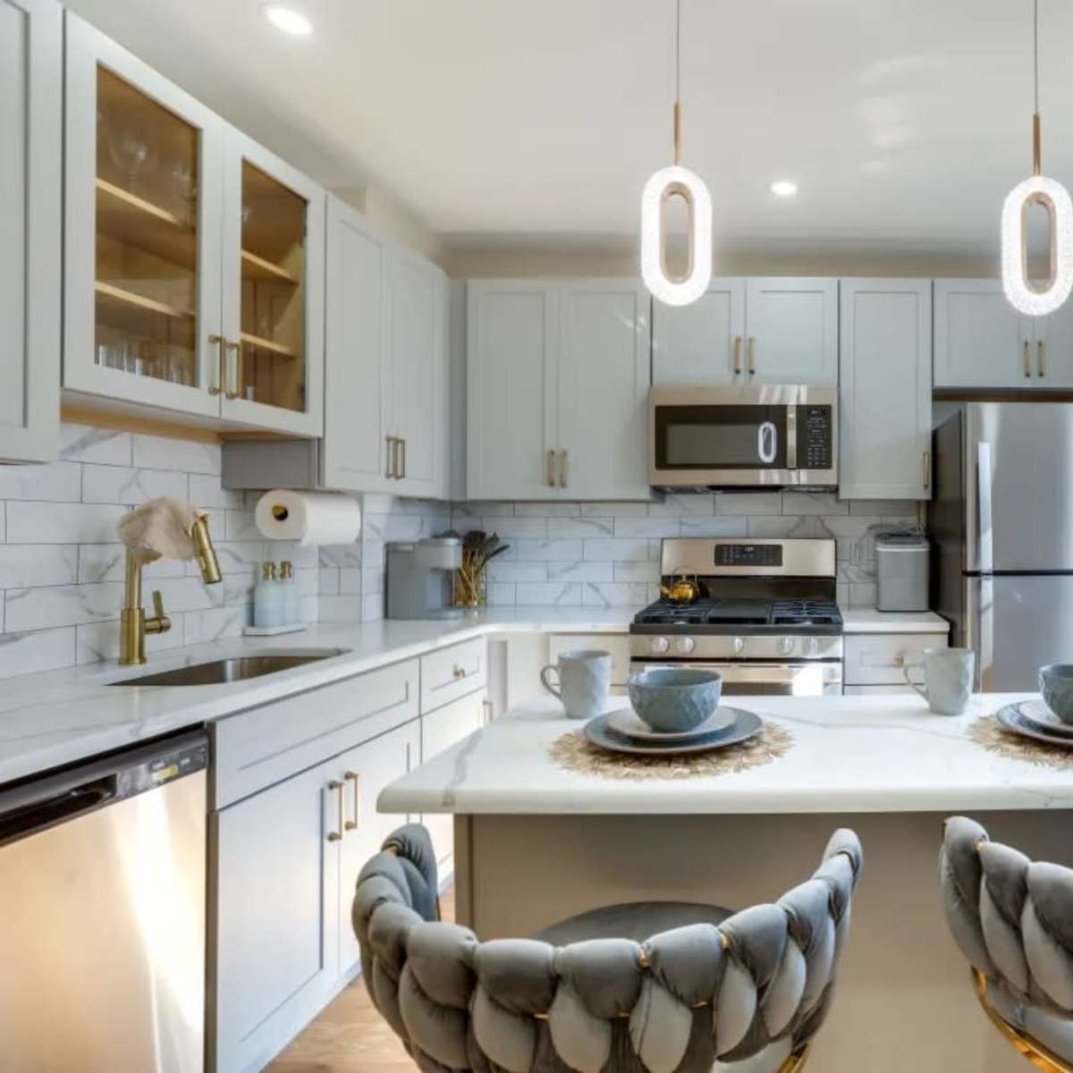 A modern kitchen features light gray cabinets, stainless steel appliances, and a white countertop with two place settings and pendant lighting overhead.