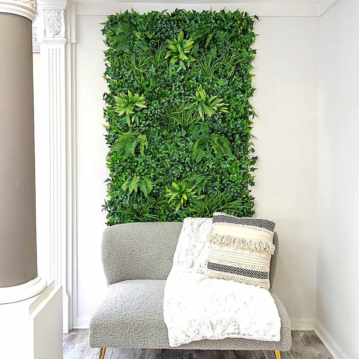 A gray sofa with a patterned throw pillow is placed against a green wall of artificial plants in a well-lit interior space.