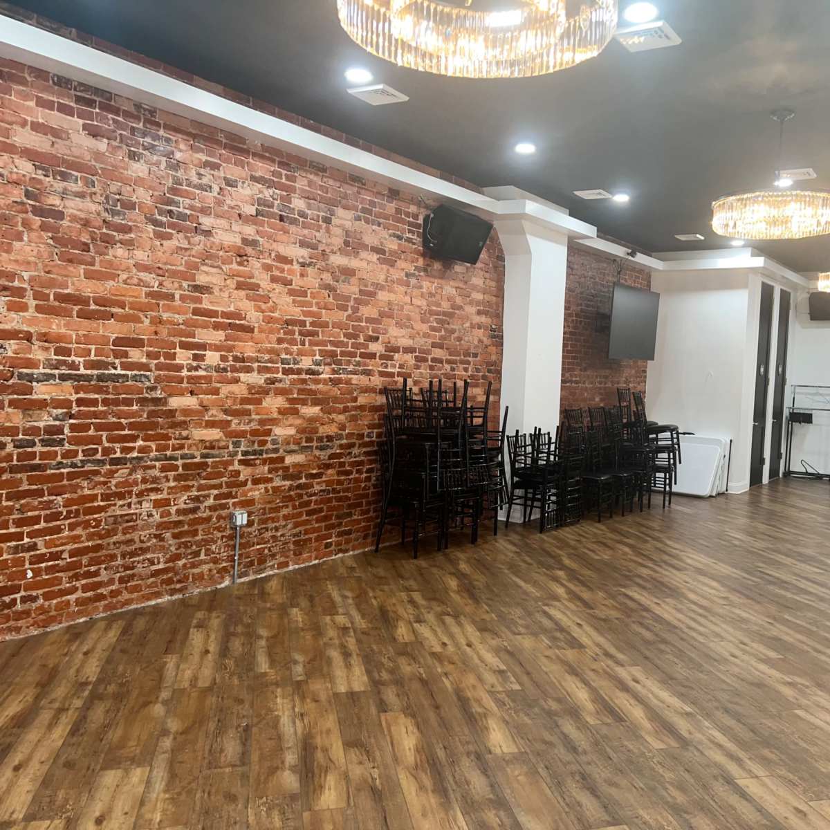 An empty room with a brick wall, wooden flooring, and stacked black chairs under modern chandeliers.
