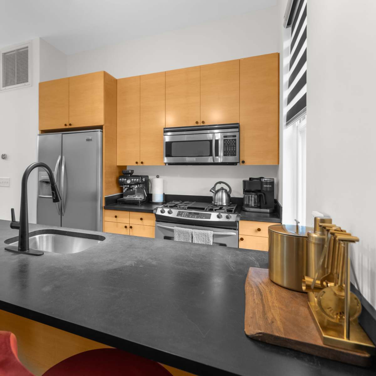 A modern kitchen features wooden cabinets, stainless steel appliances, and a black countertop with a sink and red bar stools.
