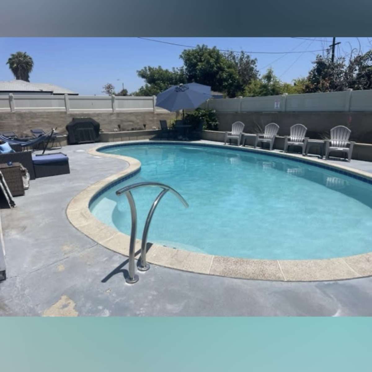 A rectangular swimming pool surrounded by patio chairs and an umbrella in a sunny outdoor area.