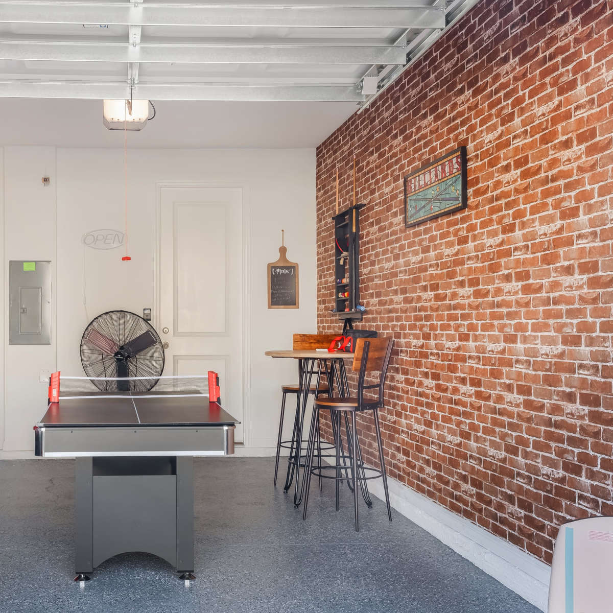 A game room with a ping pong table, arcade game, and a brick wall accent, featuring seating and a large wall fan.