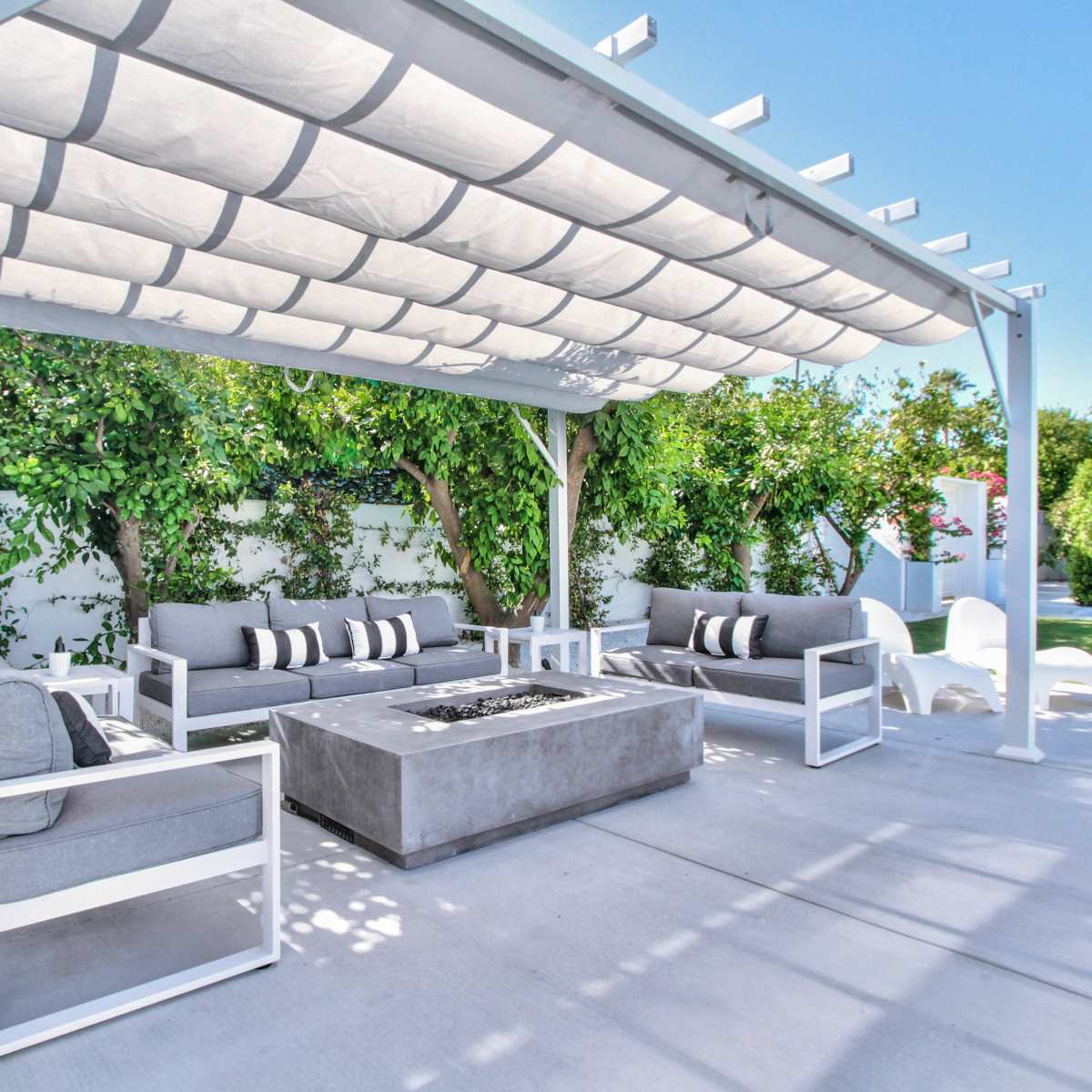A modern outdoor seating area features a fire pit surrounded by gray couches and a pergola with a canopy, set in a landscaped garden.