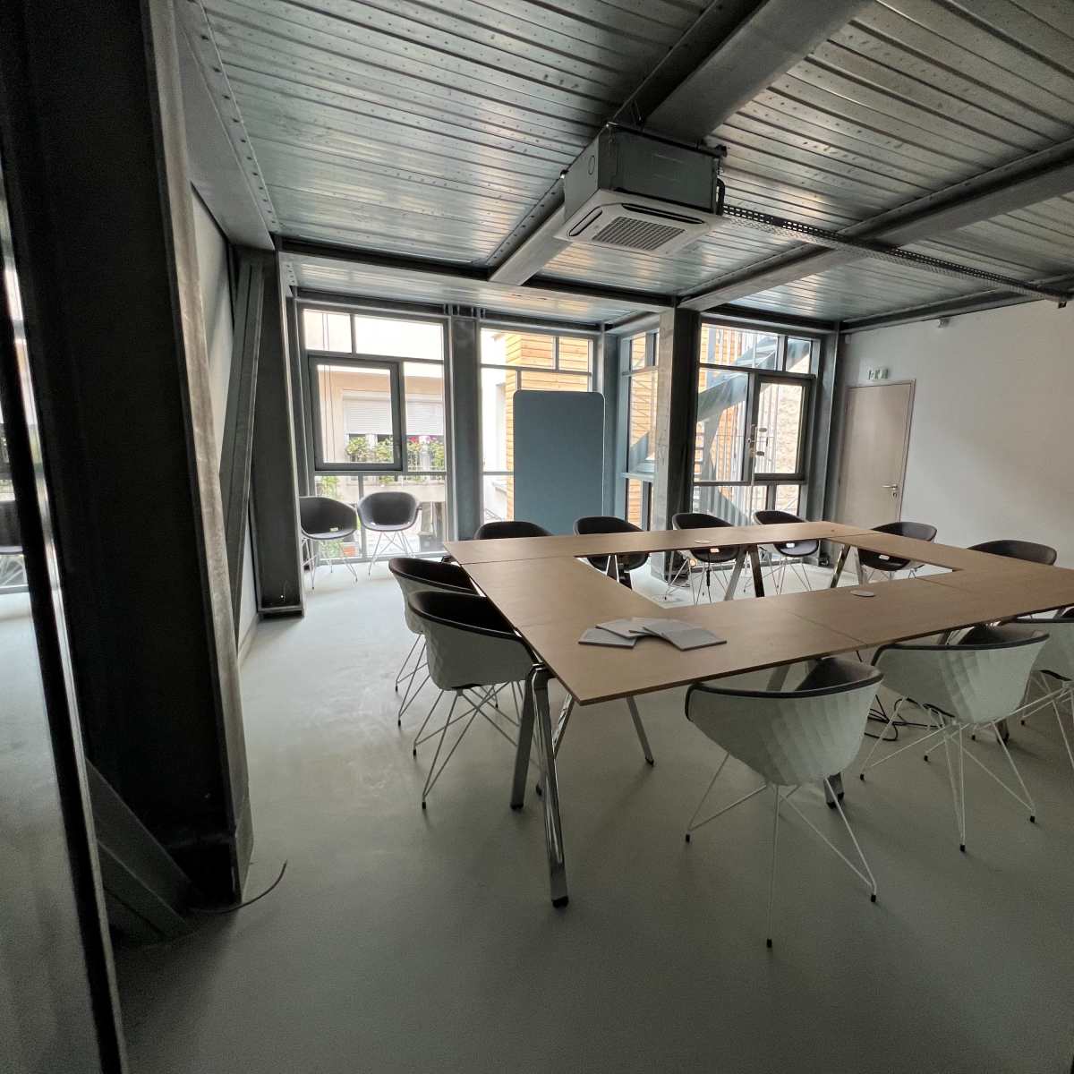 A modern conference room with a large rectangular table surrounded by white chairs, featuring windows that provide natural light.