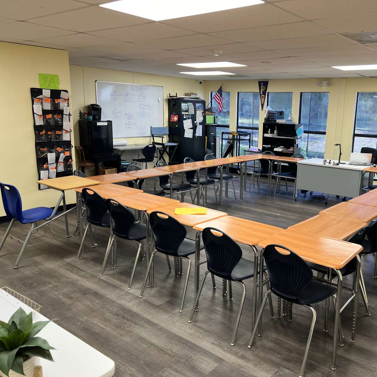 A classroom arranged with several tables in a U-shape, surrounded by black chairs, with windows and a whiteboard in the background.