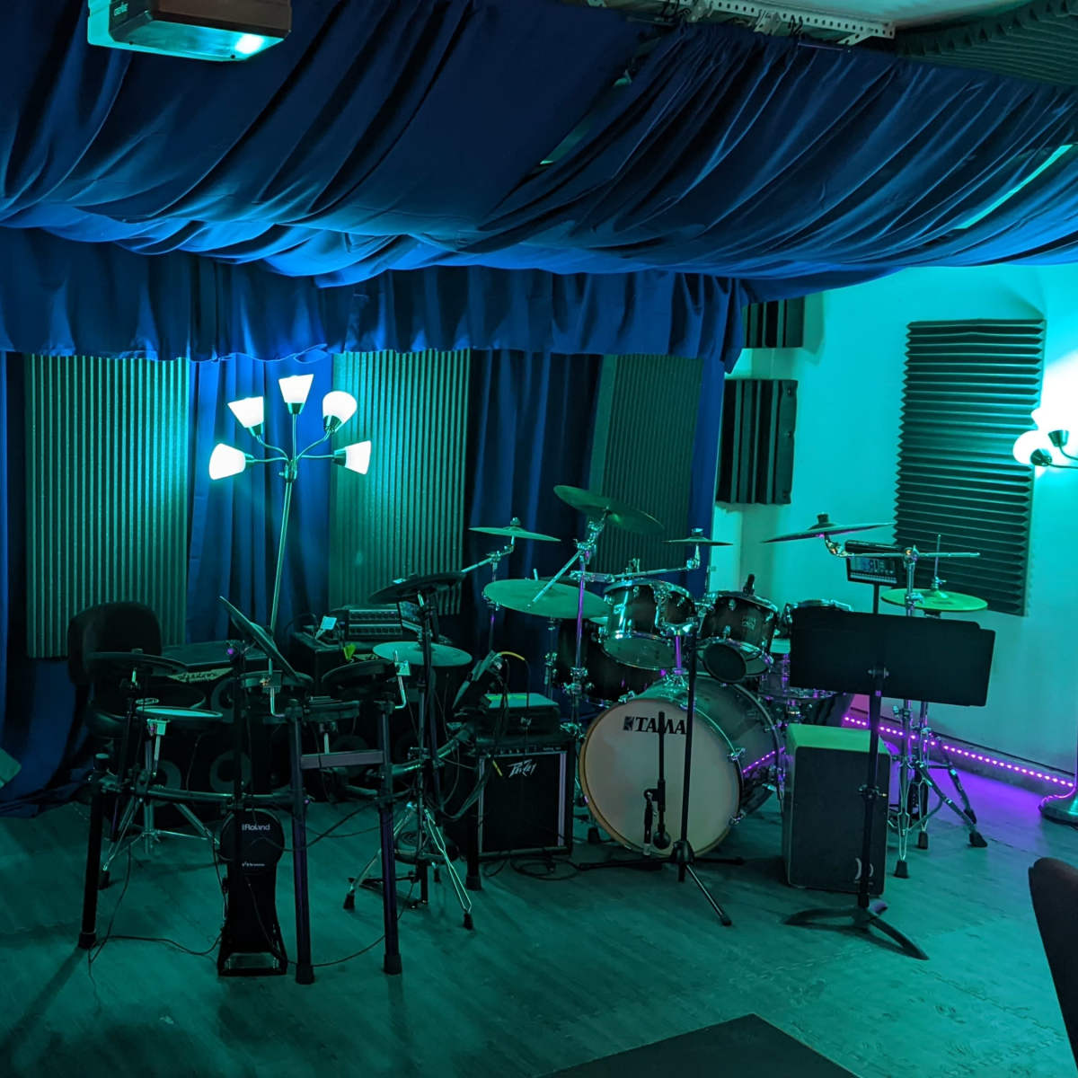 A music studio featuring a drum set, electric instruments, a piano, and various lighting elements against blue fabric drapes.