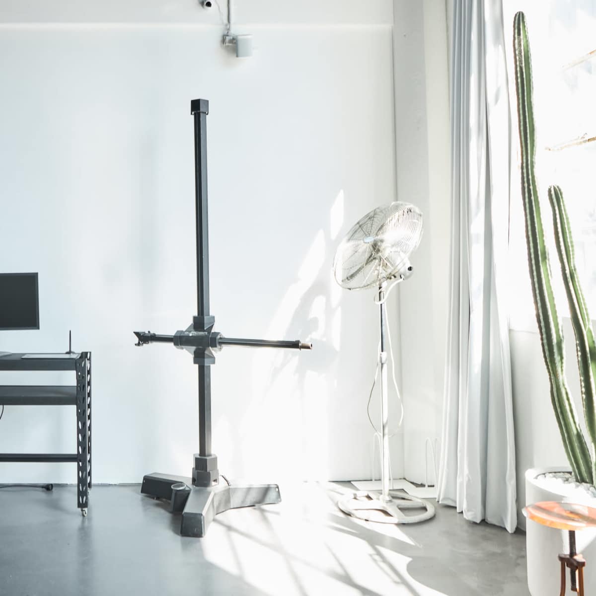A minimalistic workspace with a computer monitor on a black desk beside a tall black stand and a large fan, with a cactus in a pot nearby.