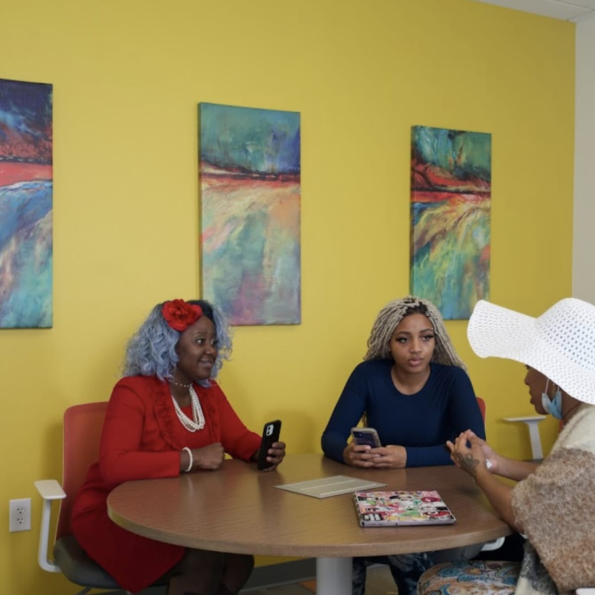 Three women sit around a circular table in a brightly colored room, engaged in conversation.