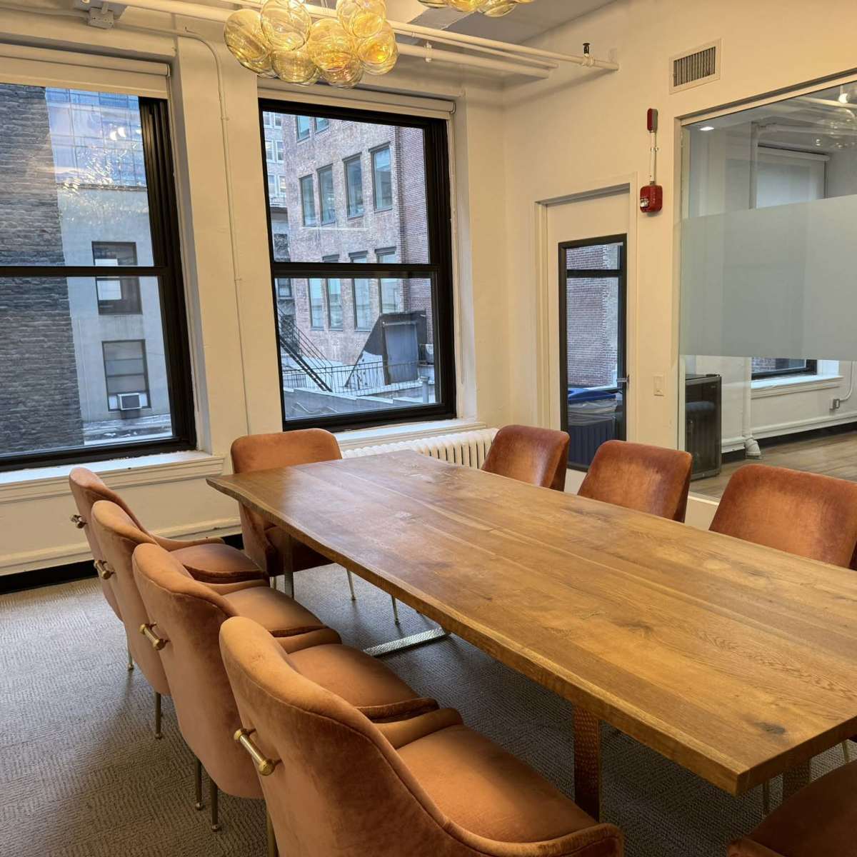 A conference room with a wooden table surrounded by upholstered chairs, large windows, and a glass wall.