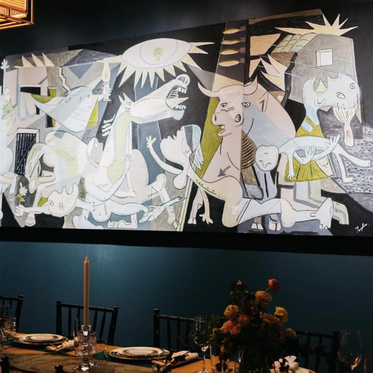 A large abstract painting hangs on a dark blue wall above a long dining table set with candles and floral arrangements.