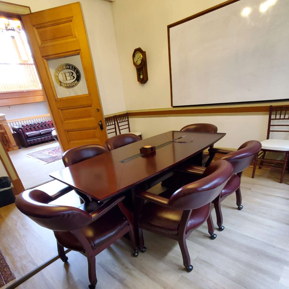 A wooden conference table surrounded by leather upholstered chairs is set up in a meeting room with a whiteboard and a clock on the wall.
