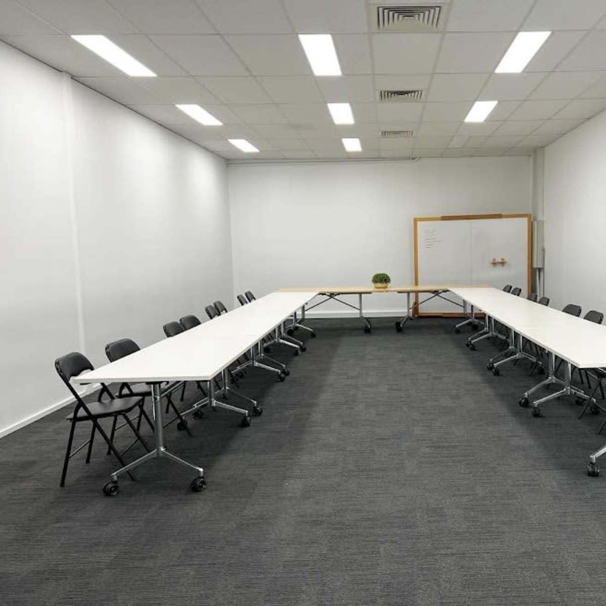 A well-lit conference room features two rows of long, white tables arranged in an elongated U-shape with black folding chairs.